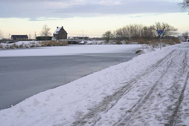 An aerial winter view shows a curved concrete dam, a snow-covered road, and a broad river winding through fields and leafless trees with a quiet, cold horizon.