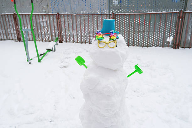 A whimsical snowman adorned with colorful decorations and a bucket hat, holding green tools in a snowy playground. Bright sunglasses add a playful touch to this winter scene.