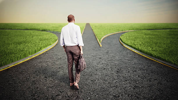 istock Man walks toward a fork in the road surrounded by green fields in the early evening light 2255441804