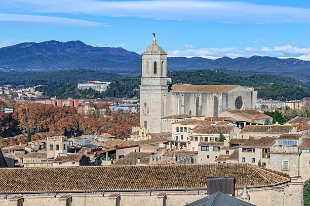 Aerial view of Girona, Spain, featuring the iconic cathedral with its distinctive bell tower dominating the historic skyline. The medieval stone buildings of the old town contrast with autumn foliage and the Pyrenees mountains creating a spectacular backdrop