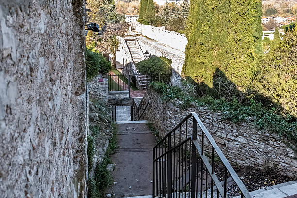 Ancient stone pathway descending between medieval walls in Parc de la Gironella, Girona, Spain, featuring metal railings and steps carved into the hillside. The historic passageway within this archaeological park is bordered by tall cypress trees and stone fortifications, offering glimpses of the surrounding residential area and autumn foliage beyond the ancient city defenses