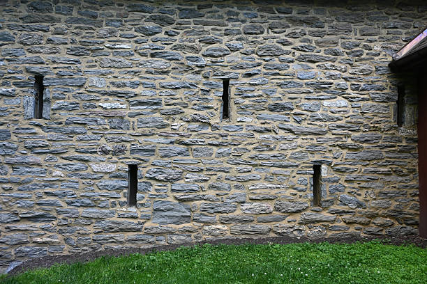 Historic Stone Wall with Narrow Vertical Window Openings, Hand-Laid Slate Masonry, and Weathered Mortar Framed by Green Grass at the Base of a Traditional Rural Building