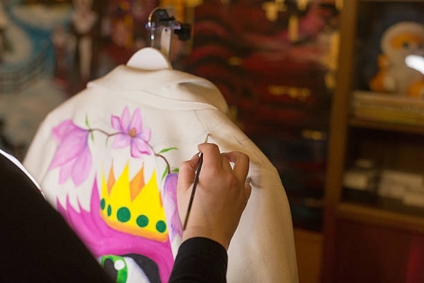Closeup artist painting jacket back, hand guiding brush over white fabric, pink floral and yellow crown motif forming, paint jars and bookshelf blurred