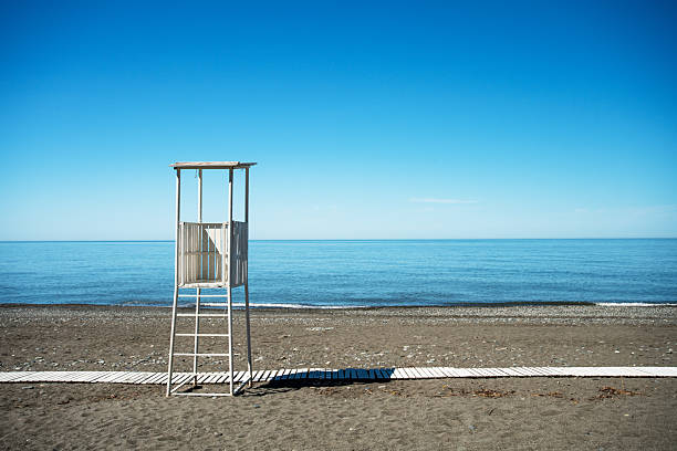 A solitary lifeguard tower sits on a calm, pebbled beach with a wooden boardwalk, facing the shimmering ocean under a bright, expansive blue sky. Tranquil, open, and timeless seaside scene.
