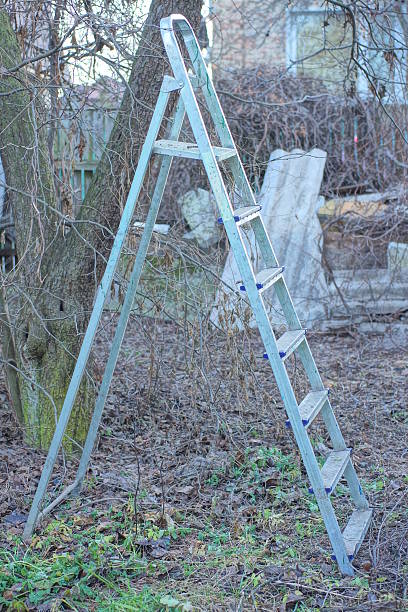 one white aluminum dirty folding ladder stepladder stands on the ground near a gray tree in an autumn garden