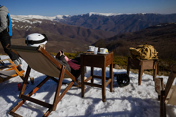 A person in ski gear enjoys a warm beverage while sitting in a deck chair on a snow-covered mountain, surrounded by breathtaking natural scenery and distant peaks