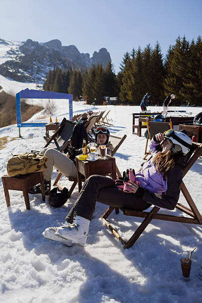 People in ski gear enjoy beverages and leisure time on a snowy mountain terrace with a scenic backdrop of pine trees and rocky peaks