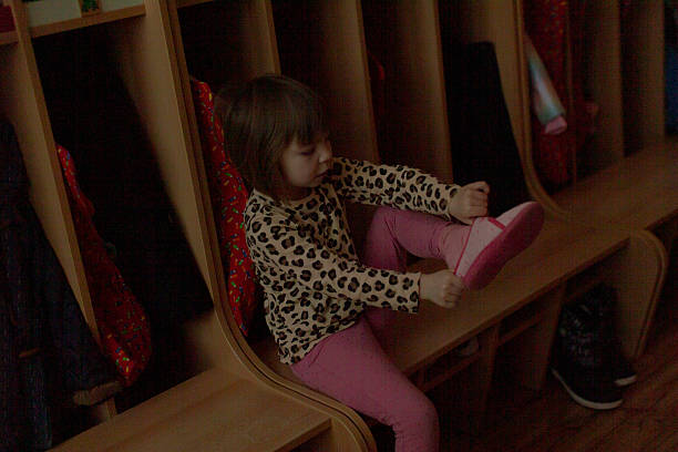 Toddler sitting in wooden cubby putting on shoe, dim hallway light and small backpack hooks, focused school readiness
