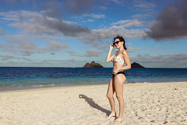 A woman in a floral bikini adjusts her sunglasses while standing on a bright, breezy Hawaiian beach, with the Mokulua Islands visible in the background.