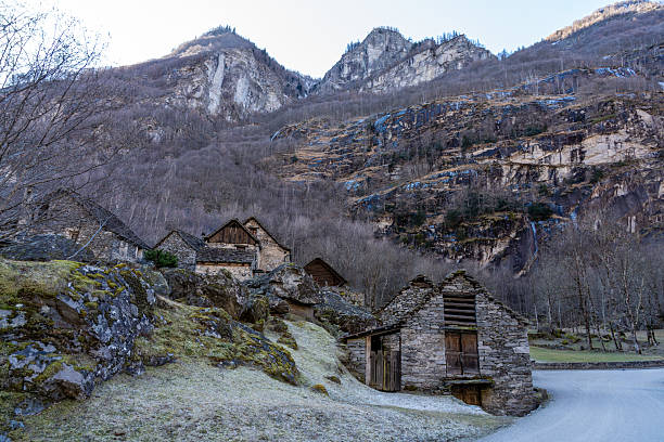 Wide shot of small stone houses (rustici) beneath a massive, steep mountain cliff.