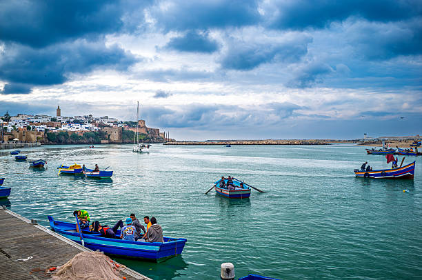 Fishing boats navigate the Bou Regreg River in Rabat, Morocco, with the Andalusian medina providing a scenic backdrop under a cloudy sky.