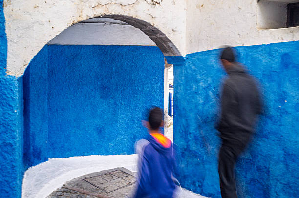 People walk through a blue and white alley in the Kasbah of the Udayas, Rabat, Morocco, showcasing the typical architecture in motion.