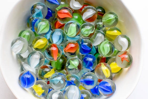 Colorful glass marbles inside a white bowl on a white table, top view flat lay, playful and nostalgic still life with clean minimal background.