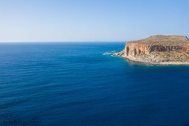 A rugged cliffside headland rises above deep blue waters at Balos Beach in Crete, Greece, with clear skies and expansive open sea creating a sense of freedom and vastness.