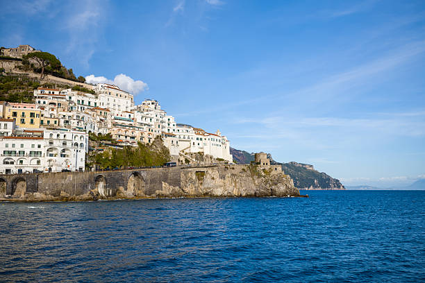 Stacked whitewashed buildings rise above stone arches along the dramatic coastline of Amalfi, Italy. An ancient stone ruin sits on a rocky outcrop overlooking the deep blue Mediterranean Sea under a clear sky.