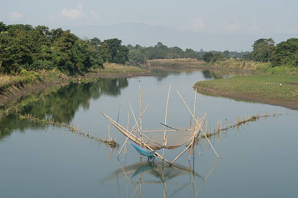 On Majuli Island in Assam, India, a traditional fishing net suspended over the river reflects the daily life of a Mising community. Supported by bamboo poles and surrounded by lush greenery, the serene scene captures the harmony between nature and ancestral fishing practices.