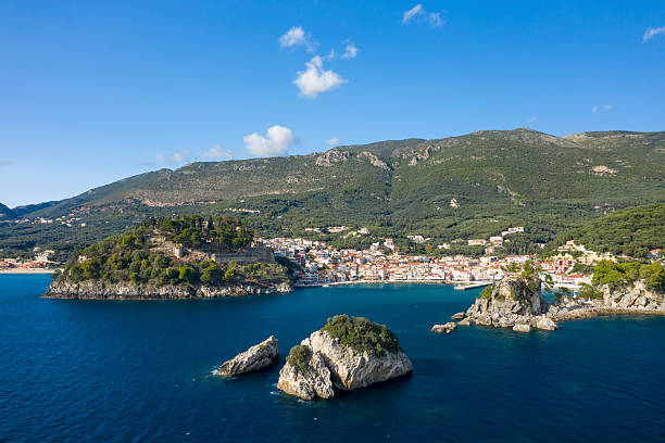Aerial view of Parga in Epirus, Greece, featuring rocky islets in deep blue Ionian waters, a historic hilltop fortress, and a colorful coastal village backed by forested hills and rugged mountains. Bright daylight and clear skies highlight the Mediterranean landscape and vibrant seascape.
