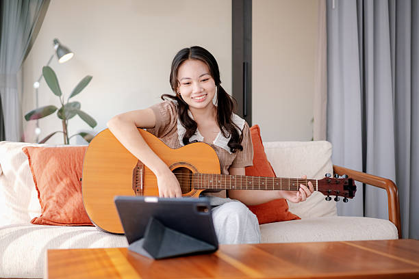 young woman sitting on a sofa at home, smiling while practicing acoustic guitar and following an online music lesson on a tablet. The scene captures a cozy and relaxed home environment