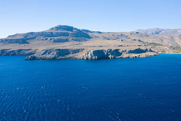 Vivid blue Mediterranean water contrasts with sunlit, rocky cliffs along the coastline near Kato Zakros, Crete. The scene features clear skies, textured stone, and an open, peaceful atmosphere.