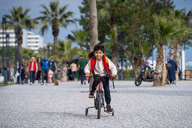 Happy young boy in a red vest and white shirt riding his bike  in a sunny park near the beach
