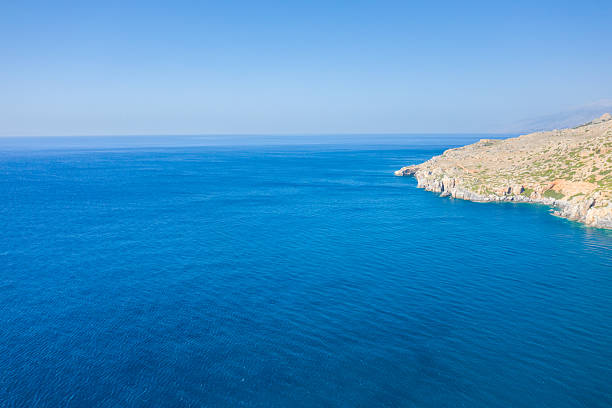 Expansive view of vibrant blue Mediterranean waters meeting a rugged, sunlit rocky coastline on the southern shore of Crete. The tranquil scene is bathed in bright daylight under a cloudless sky.