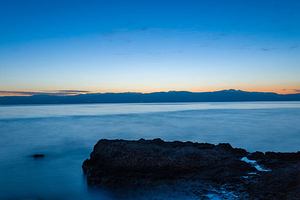 Smooth water surrounds a dark rocky outcrop under a serene twilight sky, with distant mountains silhouetted on the horizon near Kilada, Greece. The tranquil scene features soft gradients of blue and orange light.