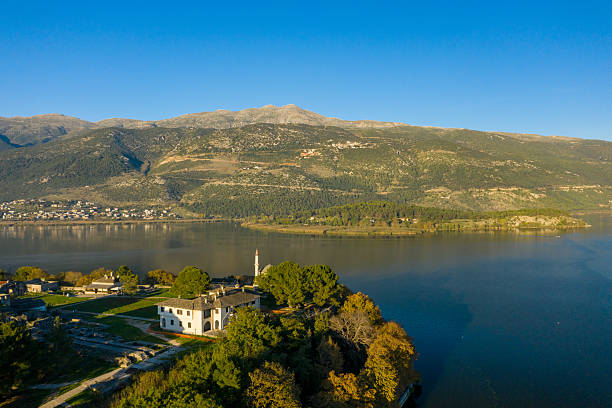 Golden morning light highlights a lakeside mosque and historic building on the edge of Lake Ioannina, with the forested slopes of the Pindus mountains rising in the background. The tranquil water and clear blue sky create a serene natural setting in Epirus, Greece.