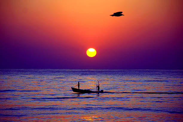 A serene ocean scene at sunset, featuring a small boat with two silhouetted figures and a bird mid-flight against a vibrant orange and purple sky. The sun is low on the horizon, casting wa