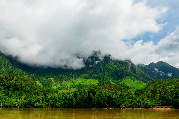 Low mist drifts across vibrant green hills and dense forest above the muddy Nam Ou River in northern Laos. The landscape features lush vegetation, dramatic clouds, and a tranquil riverside setting under a bright sky.