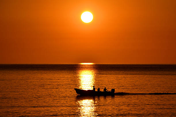 A small motorboat with three silhouetted figures glides across a calm ocean during sunset. The sun dips below the horizon, casting warm orange and red hues across the sky and water. Captured in Oman.