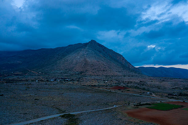 Twilight blue tones envelop a dramatic, rocky mountain rising above the arid countryside in Laconia, Greece. Low clouds and scattered farmland add depth and atmosphere to this tranquil rural landscape.