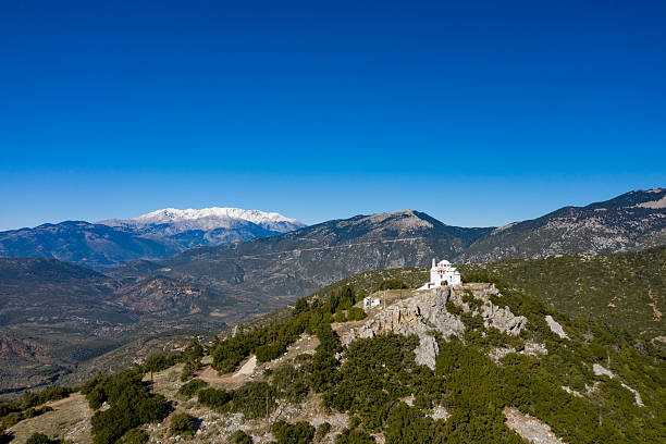 A white chapel perched on a rocky hilltop offers sweeping views of rugged green valleys and distant snow-capped peaks under a clear blue sky near Kyriaki, Central Greece. Sunlight enhances the contrast between the lush vegetation and the dramatic mountainous backdrop.