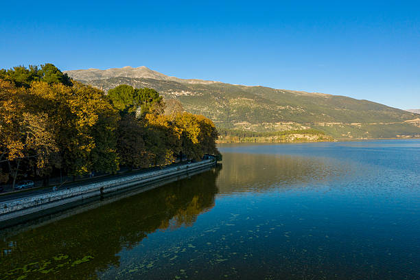 Golden autumn foliage lines the lakeside promenade of Lake Ioannina, with calm blue water reflecting the vibrant trees and distant Pindus mountains under a clear sky. The tranquil scene captures crisp daylight and natural textures in the Epirus region of Greece.