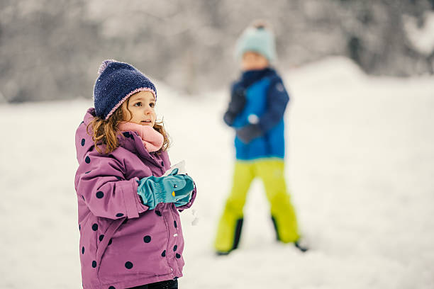 Two children laughing and playing in fresh winter snow, bundled in hats, scarves and jackets while exploring a snowy park together, enjoying playful outdoor bonding and holiday fun