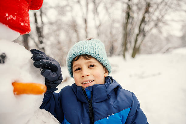 Young boy smiling in warm winter clothes building a snowman with a carrot nose in a snowy park, looking at camera and enjoying playful, carefree outdoor winter fun