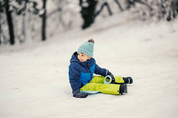 Young boy wearing winter clothing and knitted hat is happily sledding down a snow-covered hill, playing outdoors during cold weather and enjoying childhood