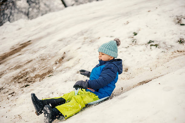 Young boy in warm hat and jacket sledding down a snowy hill, smiling with exhilaration during a bright winter day, full-length side view capturing motion and outdoor fun