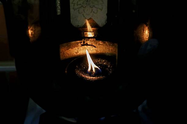 A small oil flame burns softly inside a dark metal lamp, casting warm light and shadows, creating a moody and spiritual atmosphere with a sense of ritual and calm.