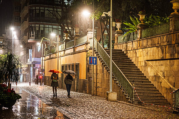 Santa Cruz, Tenerife, Spain, Europe - December 14th 2025: Two people with umbrellas hurry through a rain‑soaked night along the wet cobblestones near Parque del Príncipe. The heavy downpour creates shimmering reflections on the pavement, while the figures move quickly through the dimly lit urban scene.