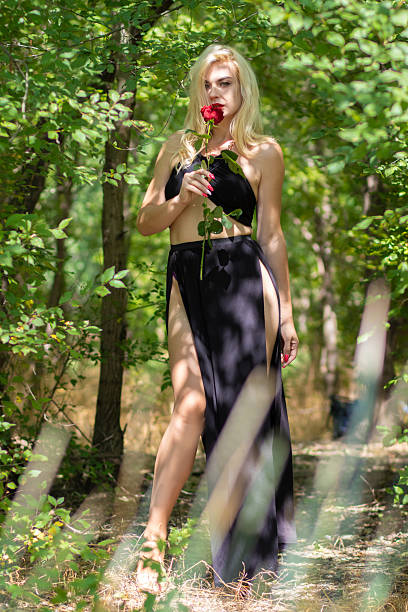 Fashion model walking in a summer forest with a red rose, black evening dress and dappled light creating a romantic and mysterious atmosphere.
