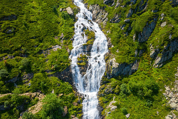 Aerial perspective of a powerful waterfall tumbling down rugged rocks surrounded by vibrant green alpine vegetation near Cascade Le Lhecou. Sunlit summer scenery highlights the dynamic water flow and textured hillside.