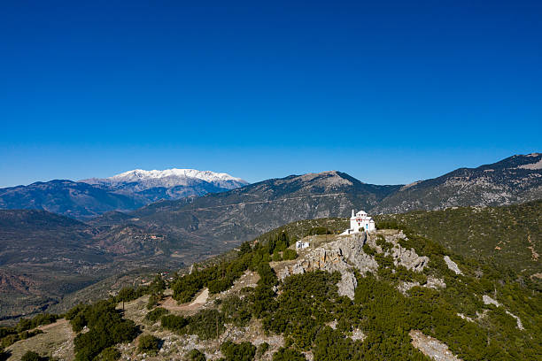 A small white chapel stands on a rocky hilltop above Kyriaki, framed by lush greenery and dramatic snow-capped mountains under a deep blue sky. The scene exudes tranquility and natural beauty with crisp winter light.