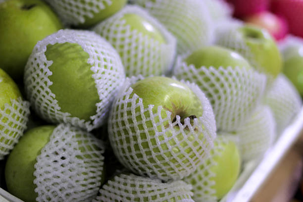Close-up of green apples individually wrapped in white foam sleeves, displayed at a grocery store. Highlights food safety, packaging, and fresh produce