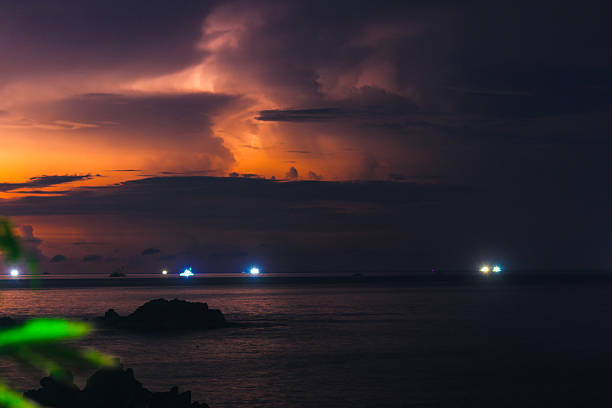 A long-exposure night seascape showing distant ship lights reflecting on calm ocean water beneath a dramatic orange-purple sky and dark storm clouds.