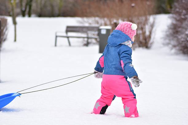 Toddler girl pulling plastic sled through fresh snow in winter park. Cute child learning balance, movement and independence during playful outdoor winter activity on a cold seasonal day.