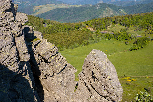 Sharp, layered basalt rock formations rise dramatically over a patchwork of meadows and dense forest, with distant rolling hills under clear summer sunlight at Mont de Gerbier de Jonc.