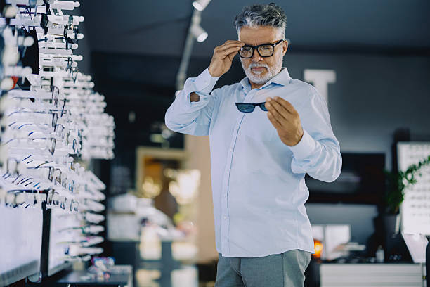 istock Senior man browsing eyeglass frames in a modern optical store. He is standing near a display wall, thoughtfully considering his options. The store has a clean, bright interior with a wide variety of eyewear on display. 2255517203