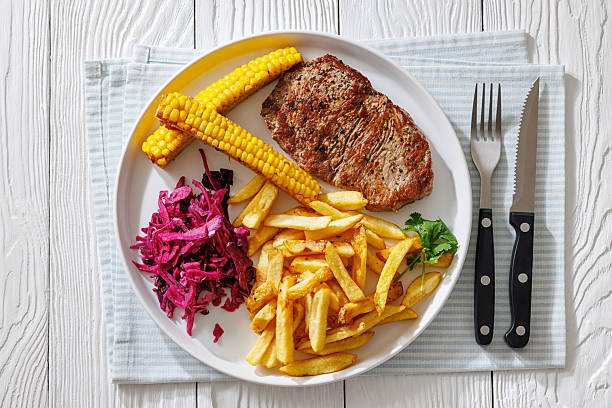 Texas-style medium rare fried beef steak served with crispy French fries, red cabbage salad, and grilled corn ribs on white plate with cutlery, flat lay, close-up, horizontal view, not AI generated
