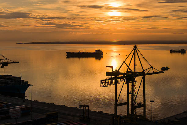 Golden sunset over a calm harbor reveals silhouette cranes, cargo ships, and reflective water. A cinematic industrial scene conveying calm, industry, and maritime commerce at dusk.