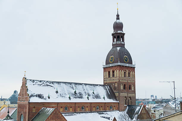 Panoramic view of Riga Cathedral featuring brick church structure clock tower and snow topped roofs representing historic religious architecture in winter city